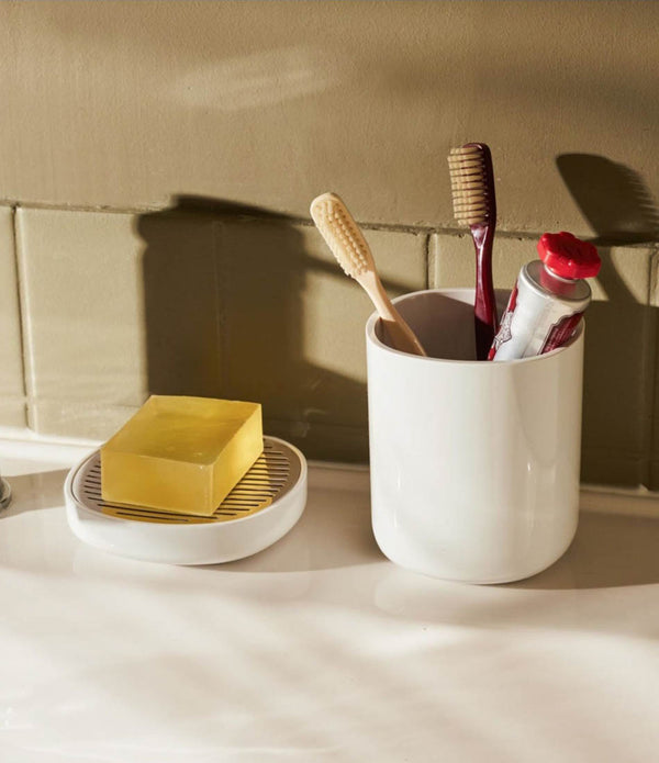 White Alessi toothbrush holder on a bathroom counter containing two toothbrushes and toothpaste, next to a soap dish.