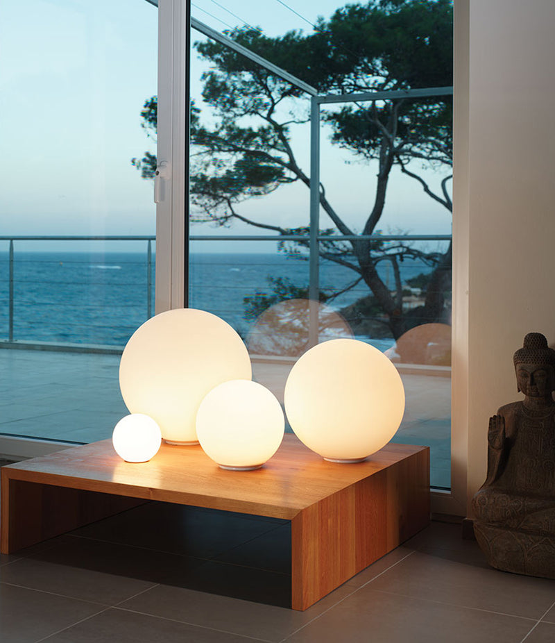 Four sizes of Artemide Dioscuri table lamps on an end table by a glass door, in a house by the sea.
