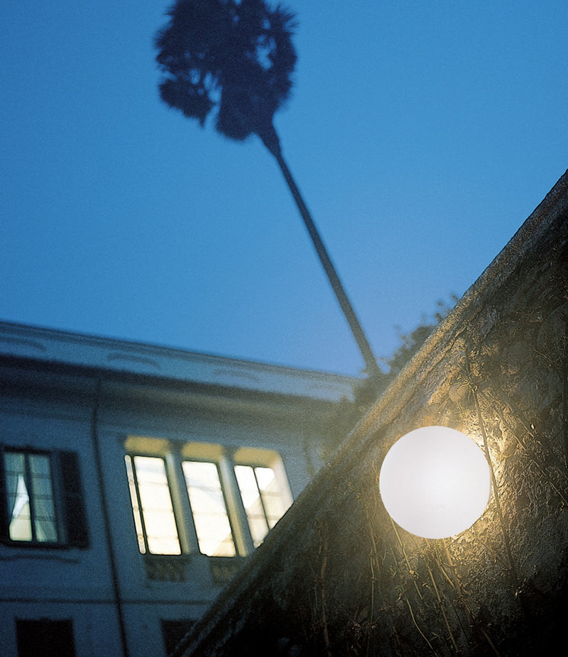 Artemide Dioscuri wall/ceiling lamp mounted to an overgrown wall beneath a palm tree.