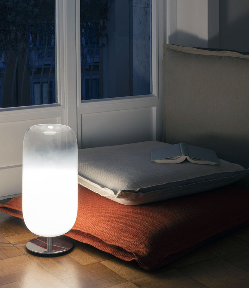 White Artemide Gople table lamp on a wood floor beside a pile of pillows.