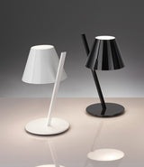 A white and a black Artemide La Petite table lamp side by side on a glass surface.