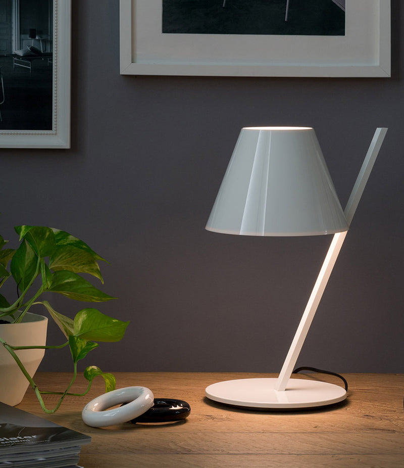 White Artemide La Petite table lamp with tilted stem and conical lampshade on a wood desk beside a plant.
