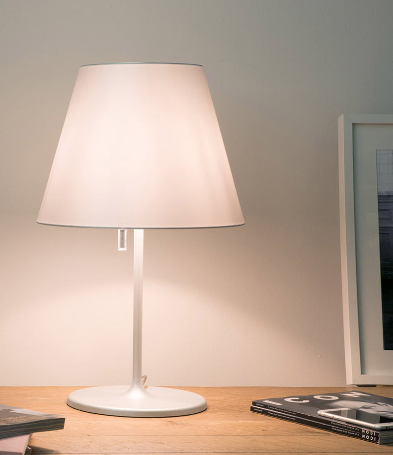 Grey Artemide Melampo table lamp on a wooden desk next to a stack of magazines.