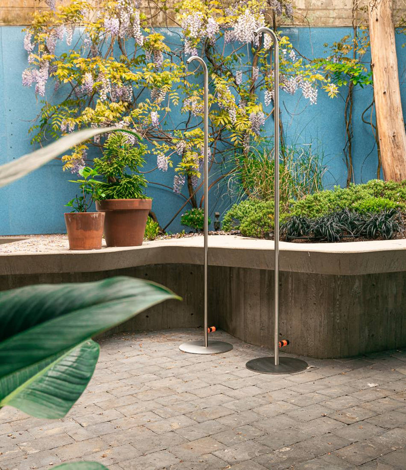 Two Coro Doccia stainless steel freestanding outdoor showers in an outdoor courtyard.