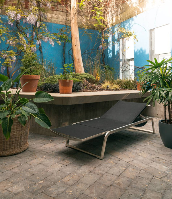 Black Coro L3 Sun Lounger in an outdoor courtyard surrounded by plants.