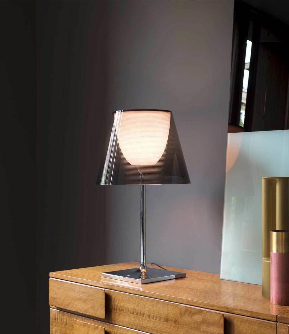 Flos KTribe table lamp on a dresser, with chrome stem and base and grey transparent lampshade.
