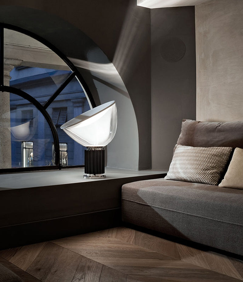 Flos Taccia table lamp with glass diffuser on a ledge beside a sofa.