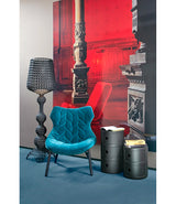 A two and three level black Componibili storage tower next to a blue chair in front of a large painting.